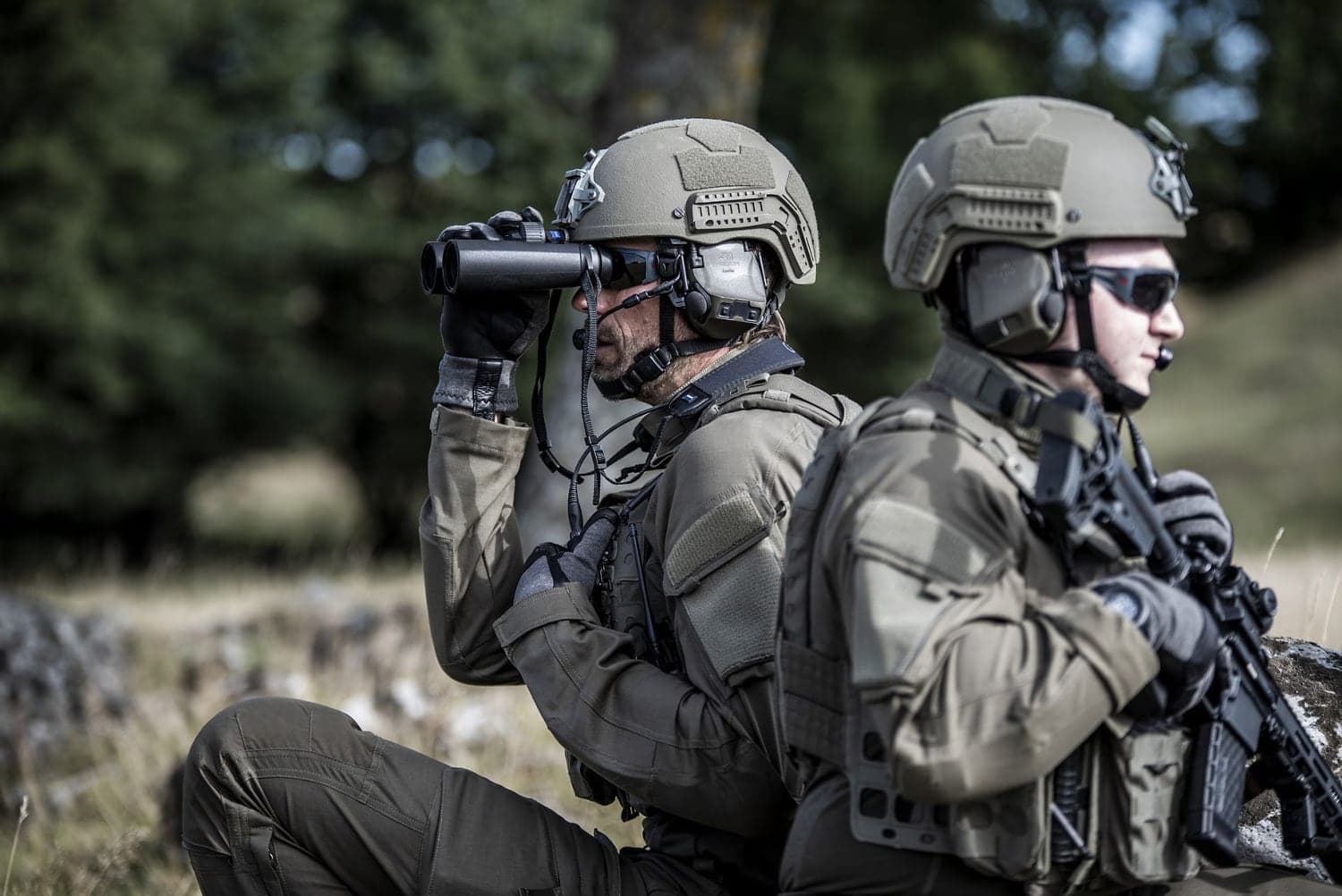 Two soldiers wearing ComTac VIII headsets under helmets, one observing with a monocular