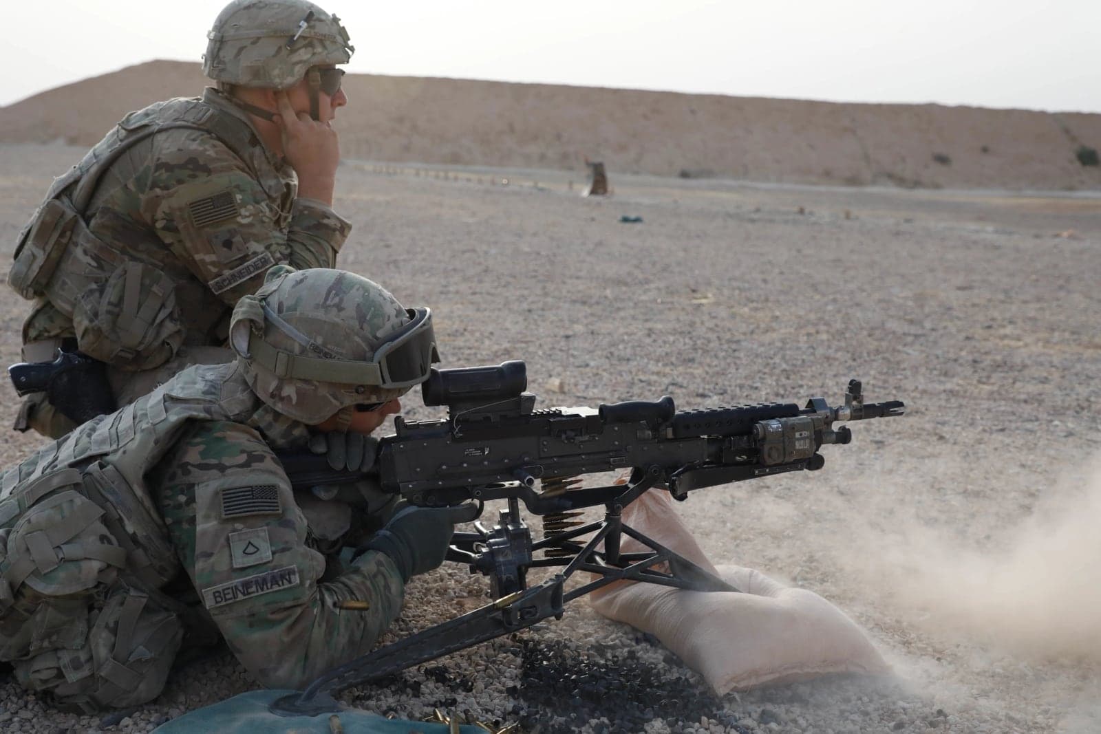 U.S. Army soldier firing an M240L from a sandbag position during a qualification range while another soldier observes
