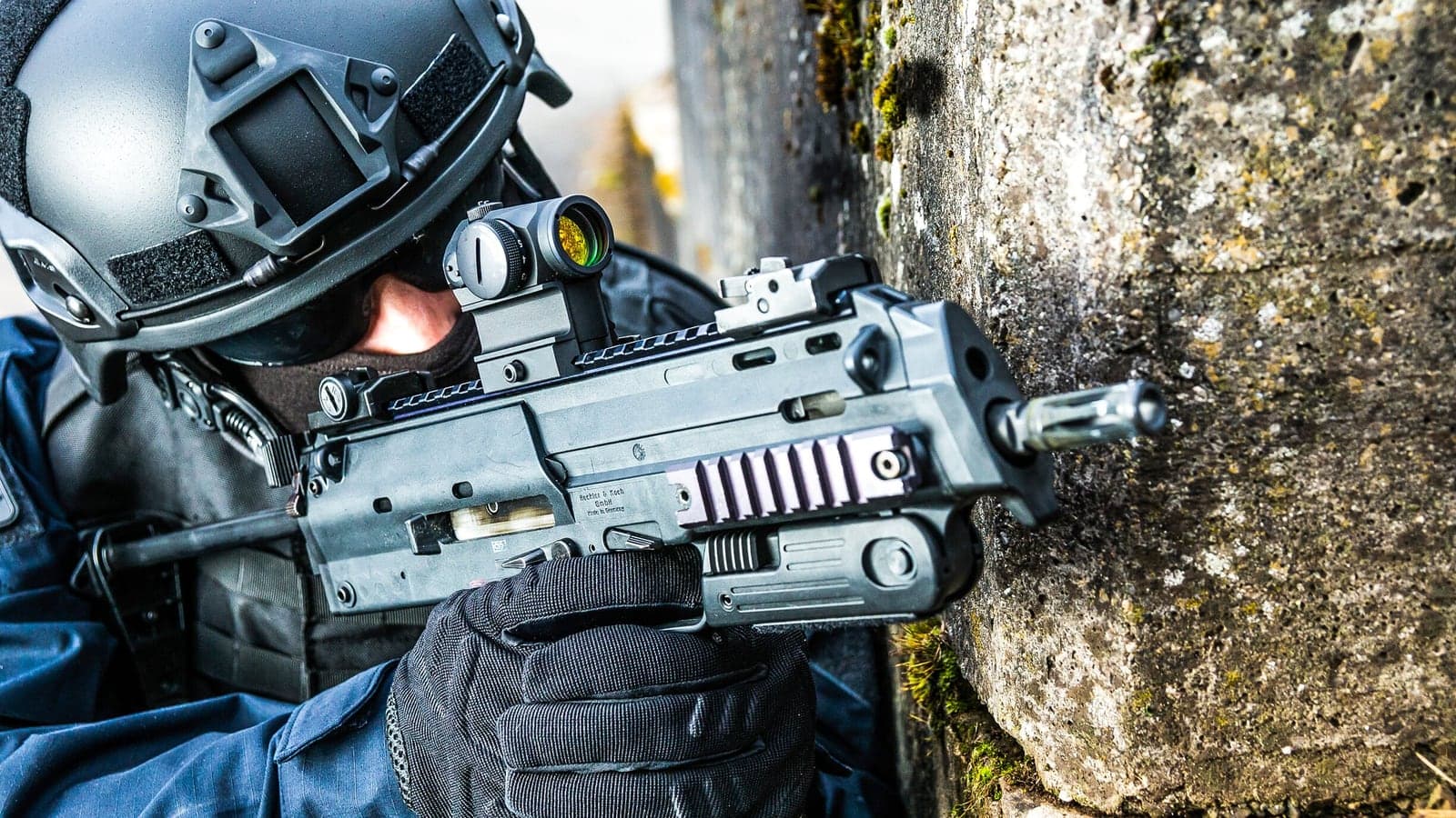 MP7 with a red-dot sight and suppressor aimed around a stone wall by a helmeted police operator