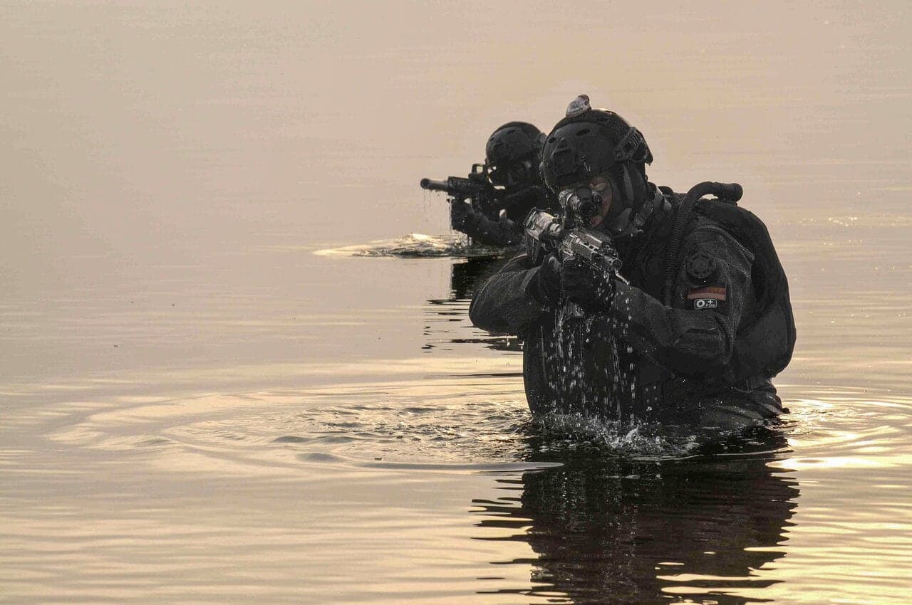 Two helmeted operators wading out of water at dusk with HK416 rifles raised