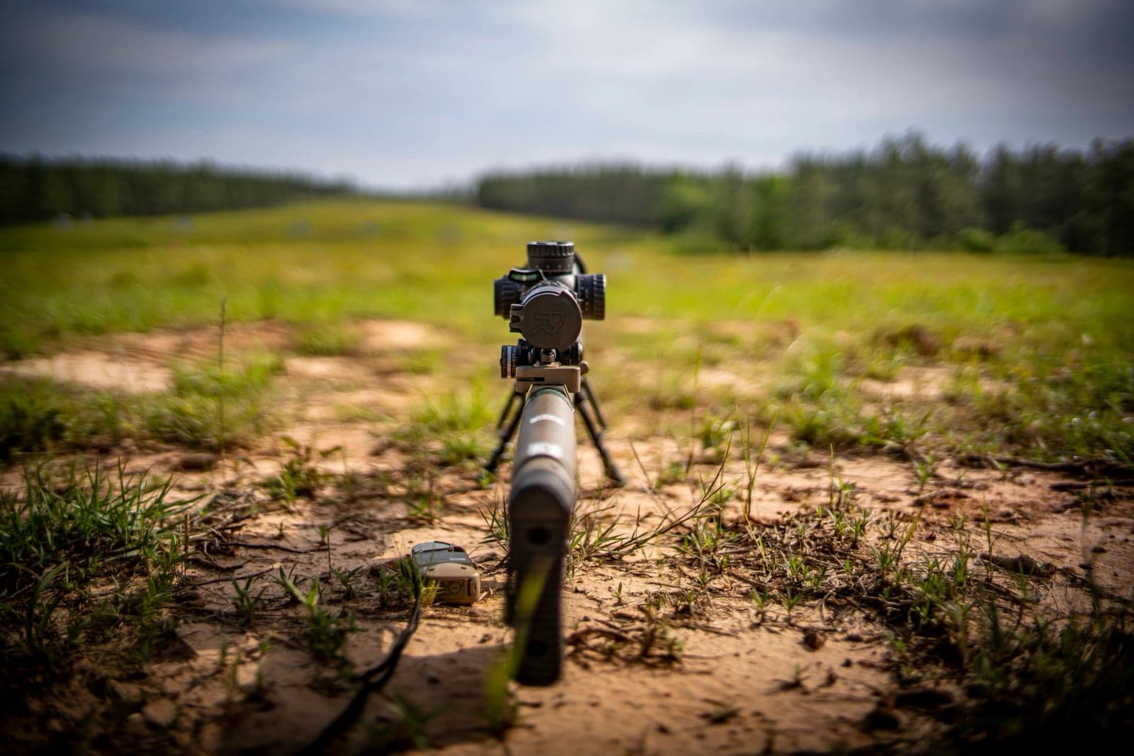 Suppressed M110 on its bipod at a grassy firing point, viewed from the muzzle end
