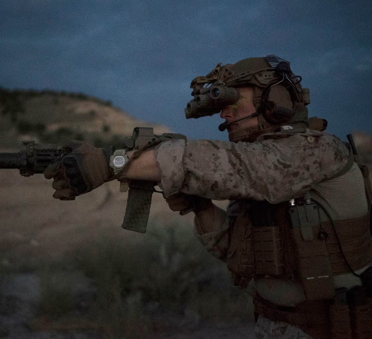 Soldier at dusk wearing a four-tube panoramic night vision goggle on his helmet while aiming a rifle