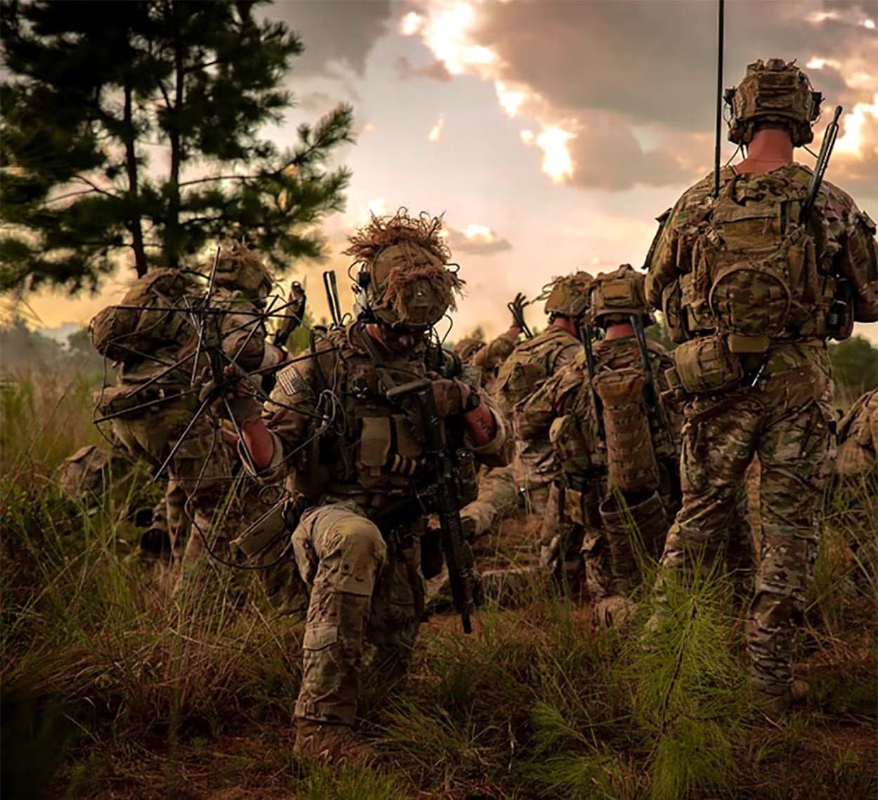 Soldiers with rucksacks and radios crouched in tall grass at dusk