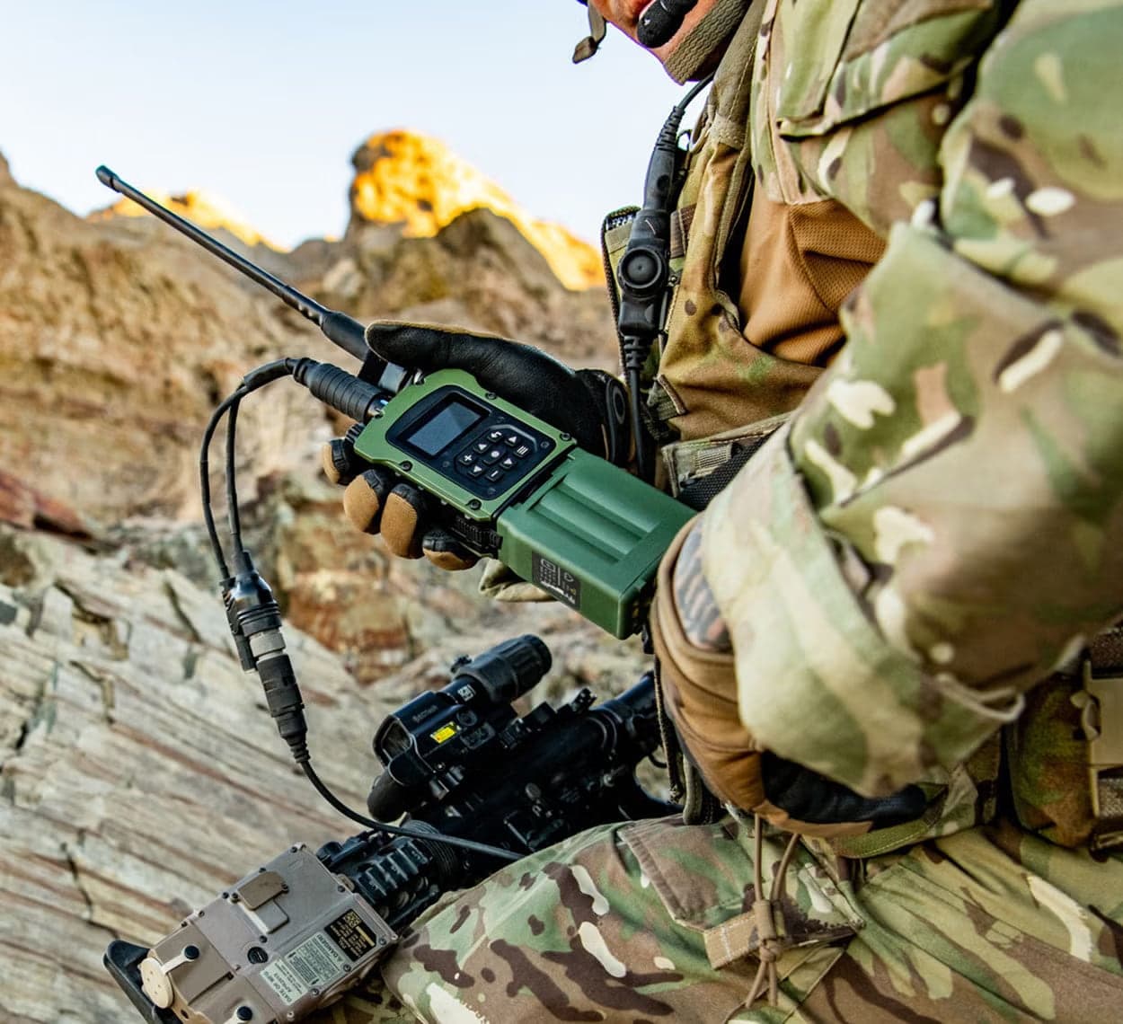 A gloved hand holding a green compact handheld radio with antenna against a rocky ridge