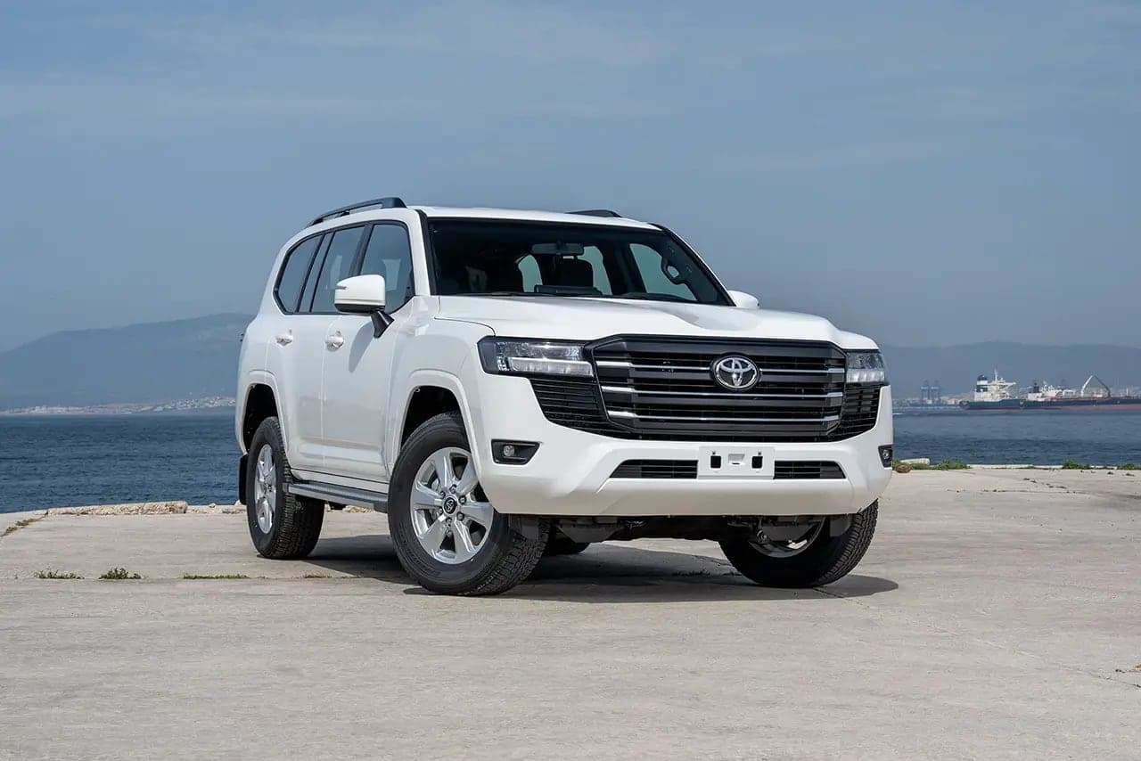 A white Land Cruiser 300 SUV parked on a concrete quay with the sea and hills behind